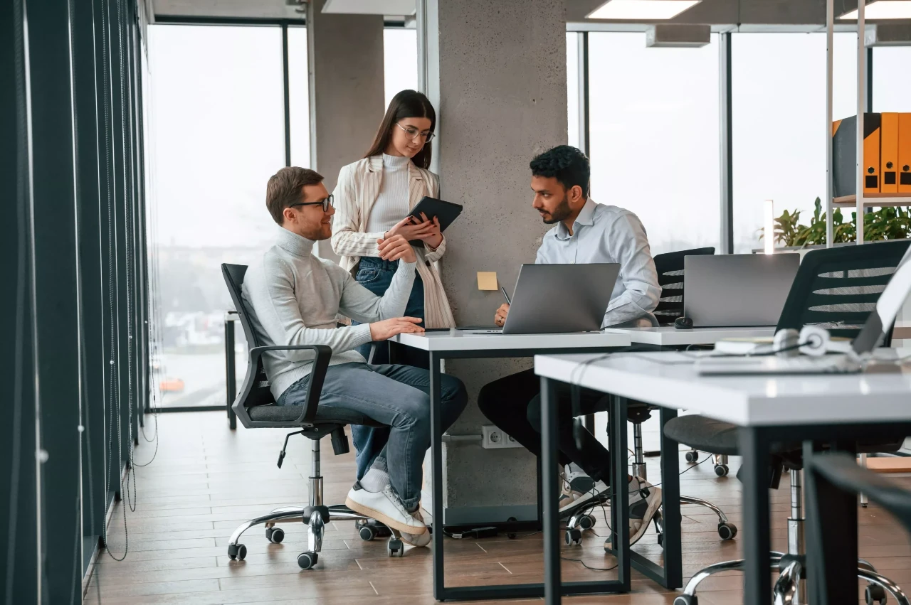 Three marketing professionals collaborating around a desk with laptops and tablets in a modern, brightly lit office space.