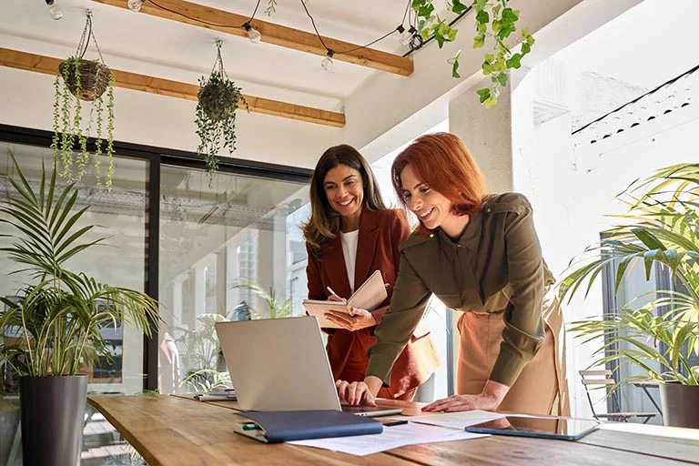 Two female colleagues smiling and working together on a laptop in a stylish, plant-filled collaborative workspace.