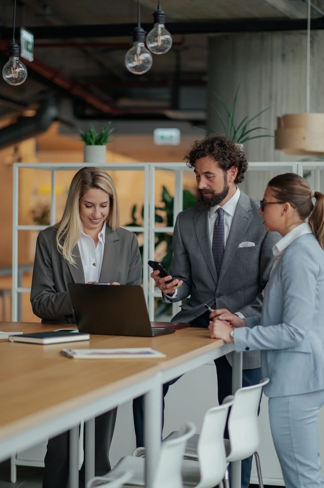 Three corporate professionals in business suits discussing a project around a high table while looking at a laptop and smartphone in a modern office.